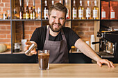 Cheerful bearded male barista in apron pouring syrup from metal jigger into glass of fresh iced coffee at counter