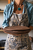 Crop unrecognizable female artisan in casual outfit and apron holding stack of flat clay plates while working in art studio