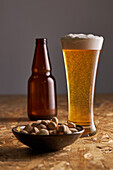 Glass of fresh cold beer placed on wooden table near bottle and plate with pistachios