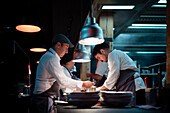 Young cooks men in aprons carefully serving meal in white ceramic dishes on restaurant kitchen