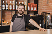 Bärtiger positiver männlicher Barista in Schürze steht neben einem Gerät und Regalen mit Glasflaschen in einem Kaffeehaus und schaut in die Kamera