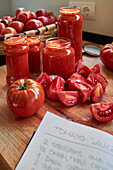 Ripe red tomatoes and jars of sauce placed on table near notebook with recipe in kitchen