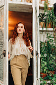 Charming young female with long ginger hair opening door of indoor garden with various potted houseplants on shelves