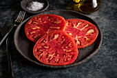 Delicious sliced tomatoes in cast iron plate near sea salt and jug of olive oil on concrete table