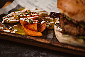 Nahaufnahme eines leckeren Burgers mit vegetarischer Frikadelle und gegrillten Shiitakes zwischen Brötchen neben Süßkartoffel- und Karottenscheiben mit Alioli-Sauce auf dunklem Hintergrund