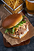 Appetizing burger with pulled pork with barbecue sauce cheddar cheese and lettuce mix served with French fries and glass of cold beer on wooden board