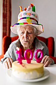 Elderly female in funny hat blowing burning candles with number 100 on birthday cake and celebrating anniversary at home