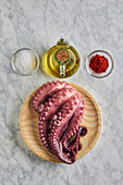 Draufsicht auf frischen rohen Oktopus auf einem runden Holzteller neben Gewürzen und Öl
