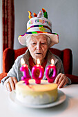 Elderly female in funny hat blowing burning candles with number 100 on birthday cake and celebrating anniversary at home