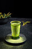 Still life composition with traditional oriental matcha tea served in glass cup with metal ornamental decor on table with ceramic bowls and fresh green leaves against black background