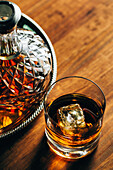 Glass of cold whiskey with ice placed on wooden table near decanter on black background