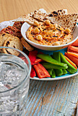 Leckerer Hummus mit Tortilla-Chips auf einem Teller mit Croutons und verschiedenen Gemüsesorten auf einem rustikalen Schneidebrett neben einem Glas kalter Limonade auf einem Holztisch mit Serviette von oben