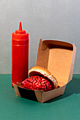 Paper box with brain burger placed near plastic bottle of ketchup on two colored background