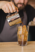 Crop bearded male barista in apron pouring ingredient from stainless pitcher into glass with ice cubes in coffee house