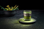 Von oben gesunder grüner Matcha-Kräutertee, serviert in einer Glastasse mit Metalldekoration auf einer mit Pulver bestreuten Untertasse auf einem schwarzen Tisch