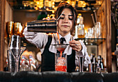 Skilled young female bartender using strainer and pouring red alcohol drink from shaker into glass with ice cubes while preparing refreshing cocktail at bar counter