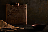 Brown hardwood cutting board in shape of square placed on table with knife and bowl in dark rustic kitchen