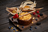 Bowl of tasty sweet cornflakes with spoon on wooden chopping board placed on table with corn cobs and berries in light kitchen