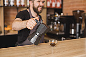 Cheerful male barista pouring hot aromatic Americano from stove top coffee maker into glass cup placed on wooden counter in cafe