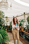 Charming female with long ginger hair standing with jug of seedling in indoor garden with various potted houseplants at home