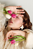 Dreamy female in dress and with delicate tulip flowers standing with closed eyes on white background in studio