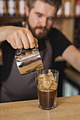 Bearded male barista in apron pouring ingredient from stainless pitcher into glass with ice cubes in coffee house