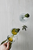 Top view of crop anonymous persons hand pouring fresh marijuana infusion into glass with herb placed on white table