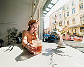 French female in beret sitting at table in cafe with aromatic glass of coffee and freshly baked croissant