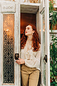 Charming young female with long ginger hair opening door of indoor garden with various potted houseplants on shelves