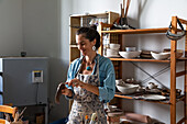 Positive adult ethnic lady in casual clothes and apron sculpting clay piece and smiling during pottery class in workshop
