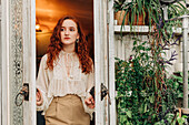 Charming young female with long ginger hair opening door of indoor garden with various potted houseplants on shelves