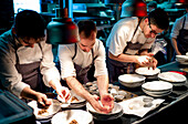 Young cooks men in aprons carefully serving meal in white ceramic dishes on restaurant kitchen