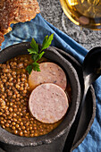 Top view rustic bowl with tasty lentil soup with parsley and slices of sausage placed on marble table and blue napkin near bread and wine during lunch