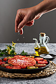 Crop anonymous person pouring sea salt on sliced fresh juicy tomato in dark bowl placed near jug of olive oil