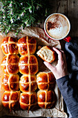 From above crop person hand holding bun on background of table with tasty lush freshly baked hot cross buns and cup of coffee