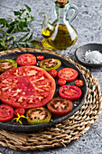 Köstlich geschnittene Tomaten in einem gusseisernen Teller auf einer Korbserviette neben einem Teller mit Meersalz und einem Krug Olivenöl auf einem Betontisch
