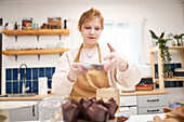 Lächelnde junge Frau mit Sieb, die Muffins in Backformen mit Puderzucker bestreut, in der Küche des Hauses