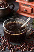 Glass of freshly brewed aromatic coffee on saucer placed on table with scattered beans against manual grinder