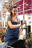 Content female seller in blue apron standing near juice making machine pouring light beverage into glass bottle