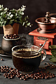 Ein Glas frisch gebrühter aromatischer Kaffee auf einer Untertasse steht auf einem Tisch mit verstreuten Bohnen vor einer manuellen Kaffeemühle