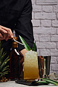 Crop unerkennbar Barkeeper garnieren Cocktail im Glas mit grünen Blättern auf Schiefer Untersetzer in der Nähe von Shaker und Flasche auf dem Tresen gegen Backsteinwand platziert