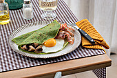 Homemade breakfast of spinach pancakes with bacon, egg and mushrooms served on a white plate with a salt and oil shaker on a checkered tablecloth