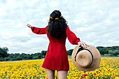 Back view anonymous trendy female in red sundress standing on blossoming field with yellow and red flowers with outstretched arms on warm summer day