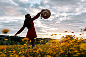 Back view of unrecognizable female with hat enjoying sundown in countryside field with blossoming flowers under cloudy sky
