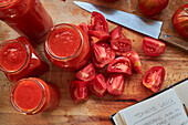 Ripe red tomatoes and jars of sauce placed on table near notebook with recipe in kitchen