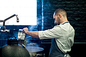 Junger Mann in grauer Schürze rührt in einer großen Metallpfanne in einer Restaurantküche