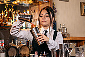 Young female bartender pouring red drink into metal shaker while preparing cocktail at bar counter