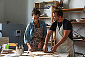 Positive middle aged female and young male in casual clothes and aprons smiling while creating clay plate during pottery class in workshop