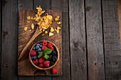 Top view of spoon with cornflakes near bowl with heap of various ripe berries on wooden chopping board in kitchen