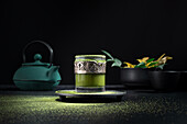 Still life composition with traditional oriental matcha tea served in glass cup with metal ornamental decor on table with ceramic bowls and fresh green leaves against black background